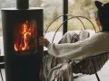 Donna avvolta in una coperta, con una tazza in mano, si scalda vicino a una stufa a legna, un modo accogliente per riscaldare la casa senza termosifoni.