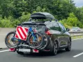 Auto in autostrada con due biciclette sul retro e box sul tetto. Il carico sporgente è ben visibile, pronto per un'avventura.