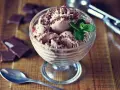 Pyszne lody nuii w pucharku, posypane czekoladą i udekorowane miętą. Obok leżą kawałki czekolady i łyżeczka.