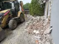 Koparka pracuje przy rozbiórce domu. Gruz i cegły leżą na ziemi, szacowany koszt rozbiórki domu jest wysoki.