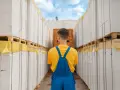 Pracownik w niebieskim ogrodniczkach stoi między rzędami palet z białymi pustakami. To obraz stabilnego systemu gospodarczego.