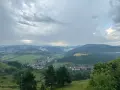 Malownicza panorama z widokiem na wieś w dolinie, otoczoną zielonymi wzgórzami i lasami. Nadciągają chmury, tworząc dramatyczne niebo nad lesnickie sedlo.