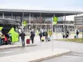 Ludzie z bagażami wsiadają do zielonego autokaru przed nowoczesnym budynkiem, przypominającym lotniska w Berlinie.
