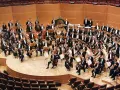 Ein großes Orchester mit vielen Musikern, die klassische Instrumente wie Violinen, Celli und Blasinstrumente spielen, posiert auf einer Bühne.