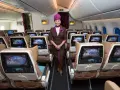 Uśmiechnięta stewardessa Etihad Airways w brązowym mundurku stoi w rzędzie foteli. Ekraniki pokazują astronautę. Dobre etihad airways opinie.
