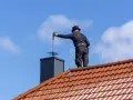 Kominiarz na dachu czyści komin. Warto wiedzieć, ile kosztuje przegląd kominiarski, by zapewnić bezpieczeństwo.