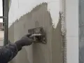 Ręka w rękawicy zatapia siatkę w tynku, tworząc wzmocnienie elewacji.