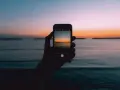 Dłoń trzyma smartfon, na ekranie którego widać piękny zachód słońca nad morzem. Idealny kadr, by nauczyć się, jak fotografować zachód słońca.