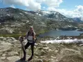 Jeune randonneuse souriante avec bâtons, face à une **mare** et des montagnes enneigées. La **difficulté** du sentier **nord** est palpable.