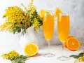 Dwa kieliszki z pomarańczowym napojem, ozdobione plasterkami pomarańczy. Obok bukiet mimozy i połówka pomarańczy. Idealne na wiosenny mimoza drink przepis.