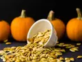 Semi di zucca freschi versati da una ciotola, con zucche arancioni sullo sfondo. Perfetti per essere piantati.
