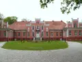 Pałac Przebendowskich w Wejherowie, różowy, z czerwonym dachem, otoczony zielenią i brukowanymi alejkami.
