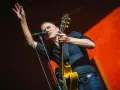 Bryan Adams na koncercie, w czarnej koszuli, z gitarą i mikrofonem, wznosi rękę w geście pozdrowienia.