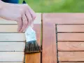 Dłoń z obrączką nakłada pędzlem brązowy preparat na drewniany blat stołu, chroniąc go przez impregnację drewna.