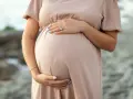 Brzuszek ciężarnej kobiety w beżowej sukience, dłonie delikatnie go obejmują. Warto pamiętać o bezpieczeństwie i unikać stosowania **retinolu w ciąży**.