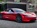Błyszczący, czerwony Tesla Roadster 2 z białymi siedzeniami stoi przed nowoczesnym domem.