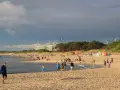 Nadmorskie miasto nad Wieprzą tętni życiem. Ludzie spacerują po plaży, bawią się w wodzie, a w tle widać budowę i wiatraki.