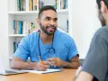 Arzt im blauen Kittel mit Stethoskop erklärt einem Patienten etwas. Ein Laptop und ein Klemmbrett liegen auf dem Tisch.