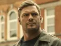 Alan Ritchson jako Jack Reacher. W tle ceglany budynek z białymi oknami.