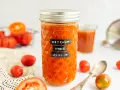 Glas mit selbstgemachtem Ketchup ohne Zucker, umgeben von frischen Tomaten. Ein Löffel liegt daneben.