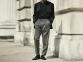 Uomo con outfit elegante: giacca scura, maglione nero e pantaloni chino grigi. Scarpe stringate lucide completano il look.