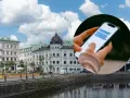 Dłoń trzymająca smartfon z widocznym oknem czatu na tle miejskiego krajobrazu z budynkami i rzeką.