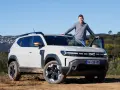 Mężczyzna opiera się o maskę jasnoszarego Dacia Duster, hybrydy z fabrycznym LPG, na tle wzgórz.