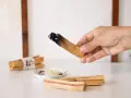 Mano sosteniendo un palo santo encendido, mostrando para qué sirve: purificación y aroma. Hay más palos y una vela encendida.