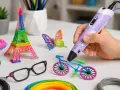 Ręka trzyma fioletowy długopis 3D, tworząc rower. Obok stoją wieża Eiffla, motyl i okulary. Widać też kolorowe filamenty, pokazujące, jak działa długopis 3D.