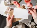 Ręce trzymają białą kartkę nad brązową kopertą. W tle prezenty i różowe tulipany, idealne na rocznice ślubu.