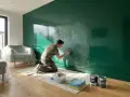 Hombre lijando pared verde brillante. Parece que la pintura no agarra en la pared, quizás por el acabado. Muebles cubiertos con plástico.