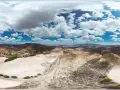 Paisaje árido con colinas, campos cultivados y un camino serpenteante. El cielo azul con nubes blancas contrasta con el desierto de larva.