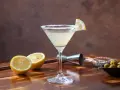 Orzeźwiający drink z martini, udekorowany plasterkiem cytryny i obsypany cukrem. Obok leżą połówki cytryny, oliwki i akcesoria barmańskie. Idealny na domowe drinki z martini.