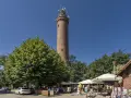 Wysoka, ceglana latarnia morska Sarbinowo góruje nad letnim kurortem z drzewami i ogródkami kawiarnianymi.