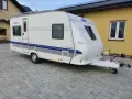 Biała przyczepa kempingowa Hobby 460 UFE "de luxe easy" stoi na kostce brukowej przed żółtym budynkiem.