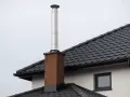 Nowy, błyszczący przedłużenie komina ze stali nierdzewnej na ceglanym postumencie, na tle ciemnego, dachówkowego dachu.