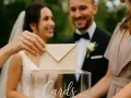 Dłoń wkłada kopertę do przezroczystej skrzynki z napisem "Cards". Młoda para w tle uśmiecha się, a goście wrzucają koperty, być może z myślą, ile na prezent ślubny.