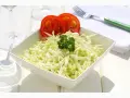 Frischer, selbstgemachter Krautsalat in einer weißen Schüssel, garniert mit Petersilie und Tomatenscheiben. Daneben liegen ein grüner Löffel und eine Serviette.