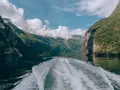 Rejs po Geirangerfjordzie. Pogoda sprzyja, chmury wiszą nad zielonymi zboczami gór, a za statkiem widać biały ślad na spokojnej wodzie.