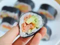 Palec wskazujący trzyma kawałek futomaki z łososiem, awokado i ogórkiem. Na talerzu widać więcej sushi.