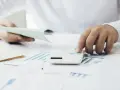 Manos calculando con calculadora blanca sobre gráficos y papeles. Una mano sostiene un talonario, la otra opera la calculadora. Proceso de factura simplificada.