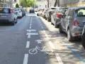 Strada a senso unico con corsia ciclabile delimitata da strisce bianche e blu. Auto parcheggiate lungo il lato destro.