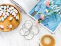 Uroczy flat lay: pastelowe drażetki, srebrna biżuteria, różowe goździki i kawa na marmurowym blacie.