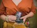 Kobieta w pomarańczowej bluzce trzyma talię kart, na wierzchu widać kartę z cyganką i napisem "3 denary tarot".