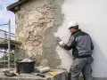 Un ouvrier en casque blanc applique de l'enduit sur une façade en cours de renovation. Échafaudage et outils à proximité.
