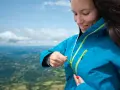 Donna con impermeabile montagna blu, chiude la zip. Paesaggio verdeggiante sotto un cielo azzurro.