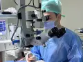 Chirurgo con maschera e camice blu opera con microscopio Zeiss. Monitor vitale e strumentazione indicano un intervento post-operatorio vitrectomia gas.