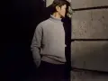 Uomo con un elegante outfit maglione collo alto grigio, in piedi vicino a un muro di pietra.