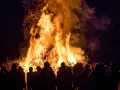 Menschen versammeln sich um ein helles Osterfeuer. Die Flammen lodern hoch in den Nachthimmel, Funken fliegen. Das Osterfeuer symbolisiert die christliche Bedeutung von Licht und Auferstehung.