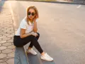Blondynka w czarnych jeansach i białej koszulce polo, siedząca na chodniku. Stylowa inspiracja, jak nosić koszulka polo damska na co dzień.